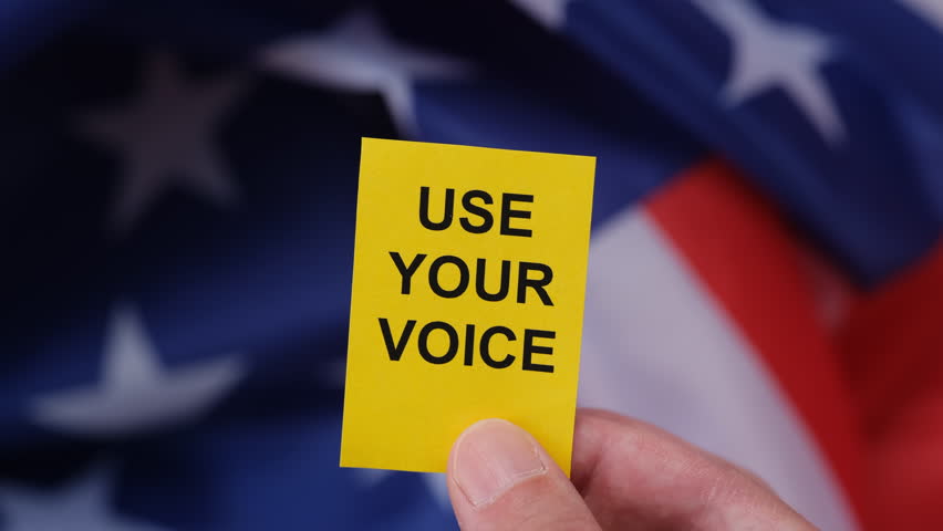 A hand holding a yellow paper note with the slogan Use Your Voice on it in front of the flag of the USA.