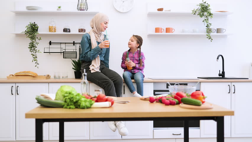 Woman in hijab sharing quality time with child surrounded by vegetables. Muslim mother and daughter drinking healthy juice together in modern kitchen setting. Family bonding over healthy lifestyle.
