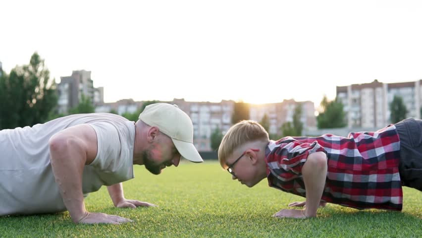 Young muscular father doing push-ups with his little son. Active happy family having fun on green grass. Fatherhood concept.