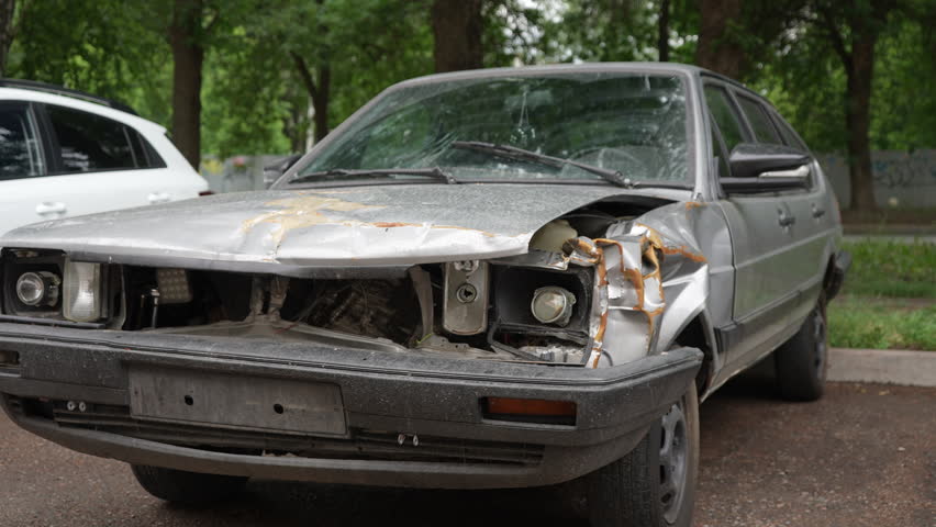 Tracking shot of destroyed abandoned car with heavily damaged front part, rusting away on side of street on cloudy summer day, evoking a sense of decay and neglect. Shooting in slow motion.