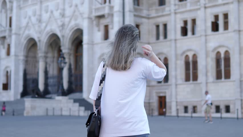 Young adult woman leisurely walks charming squares of Budapest tourist destination in Hungary. Female navigating through Budapest historical square stunning architectural details in Hungary.