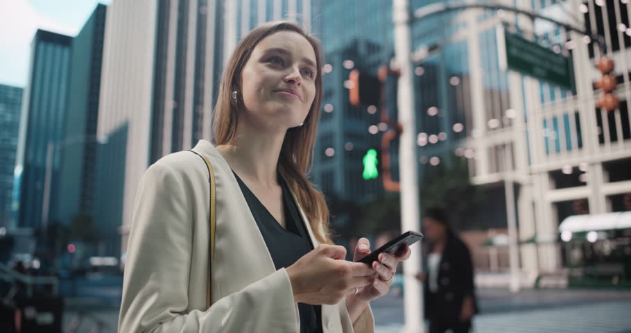 Young Caucasian Woman in a Business Outfit Using Smartphone while Standing on a Busy City Street, Surrounded by Tall Buildings and Traffic. Female Browsing Internet and Checking Social Media