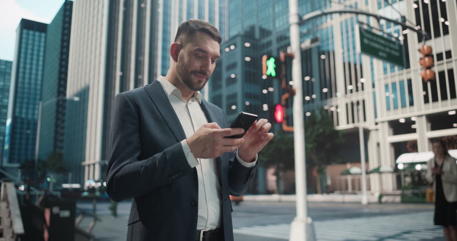 Young Caucasian Man in a Business Outfit Using Smartphone while Standing on a Busy City Street, Surrounded by Tall Buildings and Traffic. Smiling Man Browsing Internet and Checking Social Media