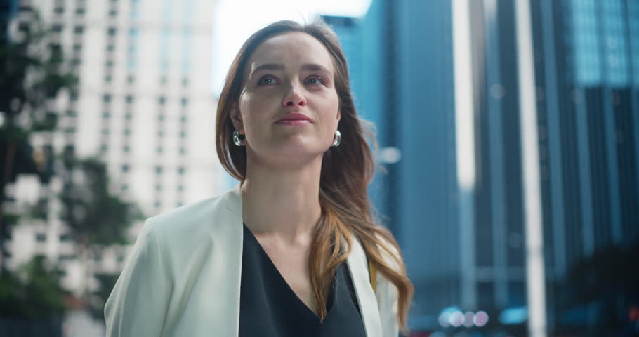 Confident Young Woman in a Stylish White Blazer Standing on a City Street, Looking Around and Positively Smiling. Background with Towering Skyscrapers, Business Buildings and Urban Neighbourhood