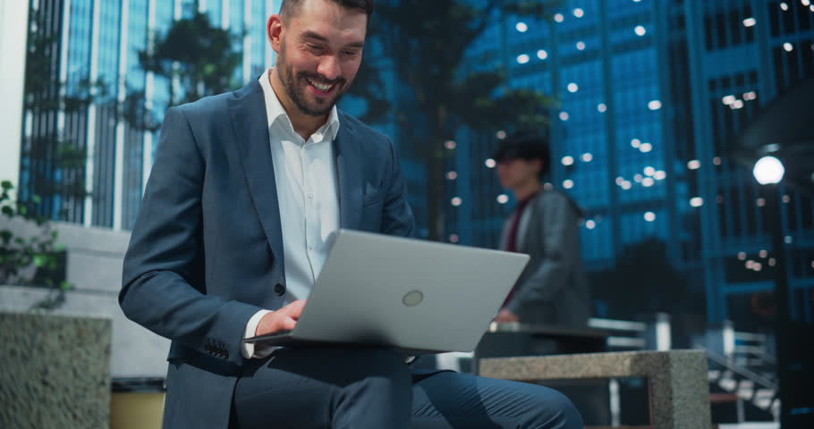 Portrait of a Cheerful Businessman in a Blue Suit Sitting in a Park in a Modern Urban Plaza, Typing on His Laptop Computer. Manager is Responding to Urgent Emails and Managing His Project Deadlines