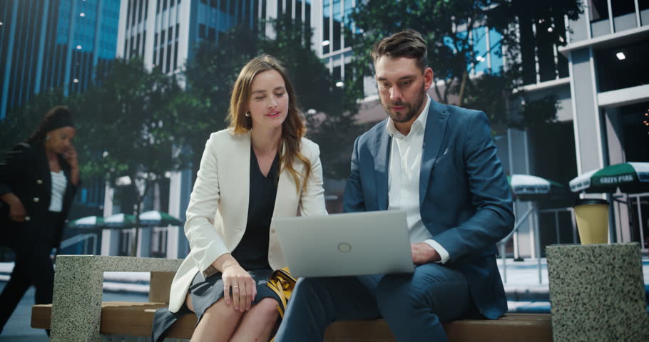 Businessman and a Businesswoman Sit Side by Side on a Park Bench in a Busy Urban Area, Both Focused on a Laptop Computer Screen. Colleagues Collaborating on a Joint Report for an Upcoming Presentation