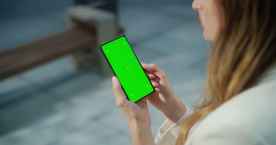 Female Holding a Green Screen Mock Up Chroma Key Smartphone on a Street. Over the Shoulder Footage of a Person Using Internet, Social Media, Online Shopping with Mobile Phone Device