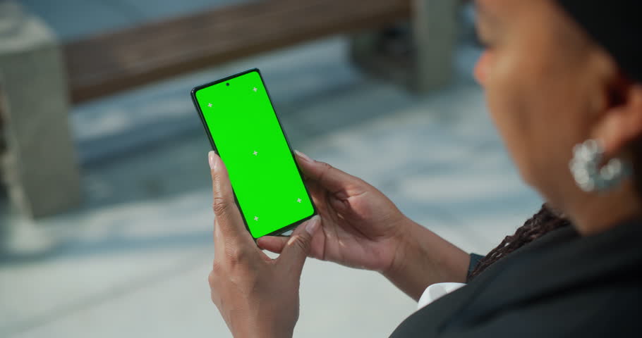 Female Hand Holding a Smartphone with Green Screen Mock Up Display. Black Woman is Standing on a Street and Checking Content on a Mobile Device. Close Up Over the Shoulder Footage