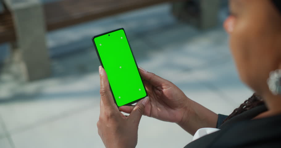 Female Holding a Green Screen Mock Up Chroma Key Smartphone on a Street. Over the Shoulder Footage of a Person Using a Mobile Phone. Template for Internet, Social Media, Online Shopping App