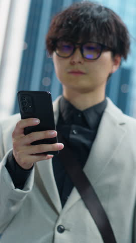 Vertical Screen: Young Asian Man Using Smartphone while Standing on a Busy City Street, Surrounded by Tall Buildings and Traffic. Tech Company Founder Browsing Internet and Checking Social Network