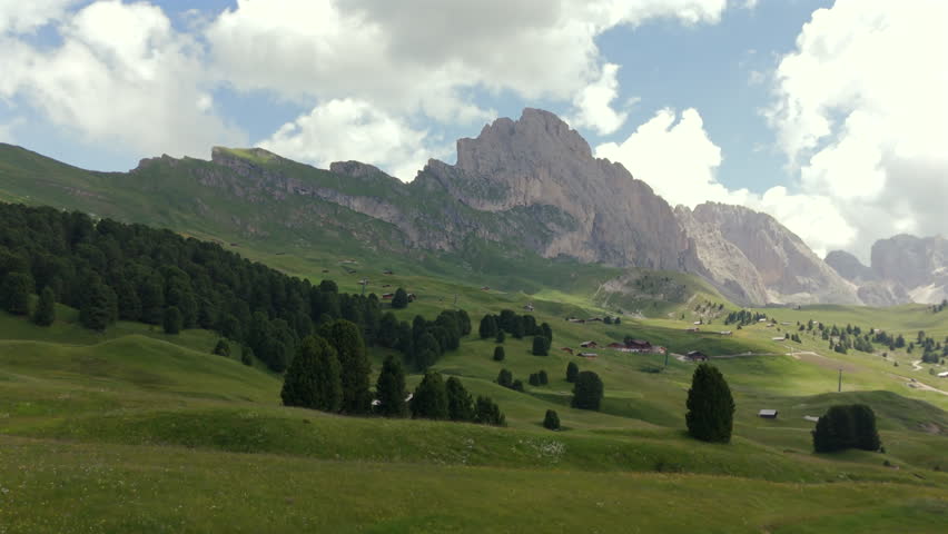 A picturesque alpine meadow with traditional wooden cabins, set against a backdrop of towering mountains and lush green forests under a cloudy sky.
