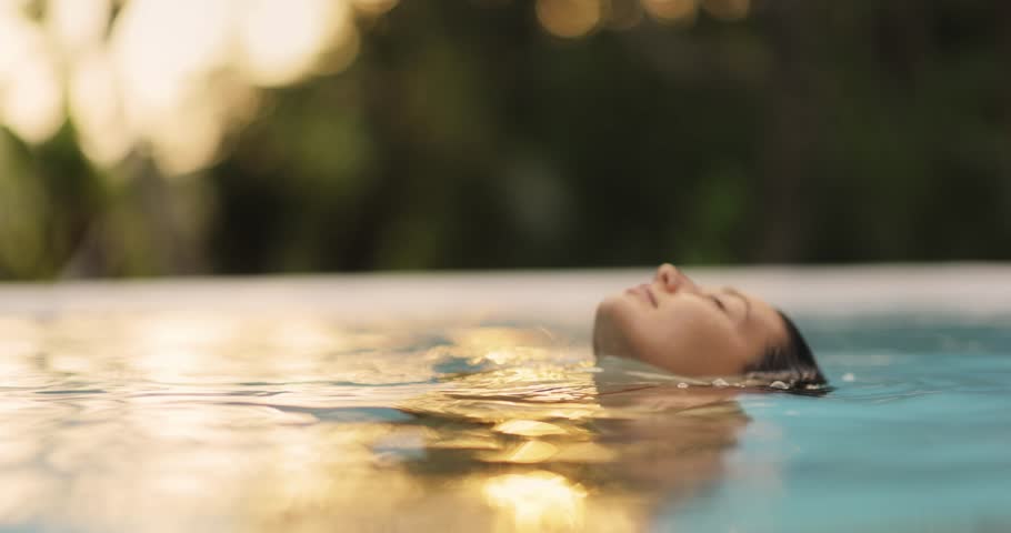 Tropical paradise, luxury relaxation of bikini brunette woman in sexy orange swimsuit in infinity pool at sunset covering her breast by hands. High quality portrait slow motion 4k footage