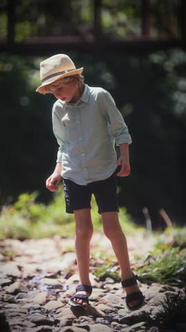 A young boy wearing a sunhat walks cautiously on a rocky path, nearly losing his balance during the daytime