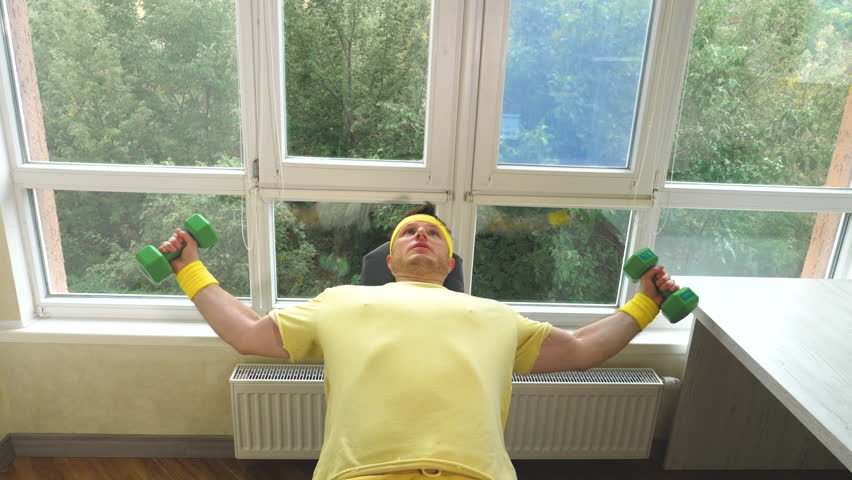 A young athlete man pumps his chest muscle with dumbbells. An athlete with a headband training on a home gym. Against the background of windows. Slow motion