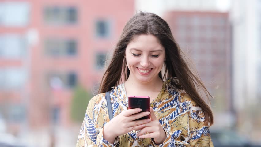 Beautiful woman with long blond hair holding mobile phone, looking at mobile phone screen, using smartphone apps. posing on street. Positive model smiling. She is standing with smartphone in hands on