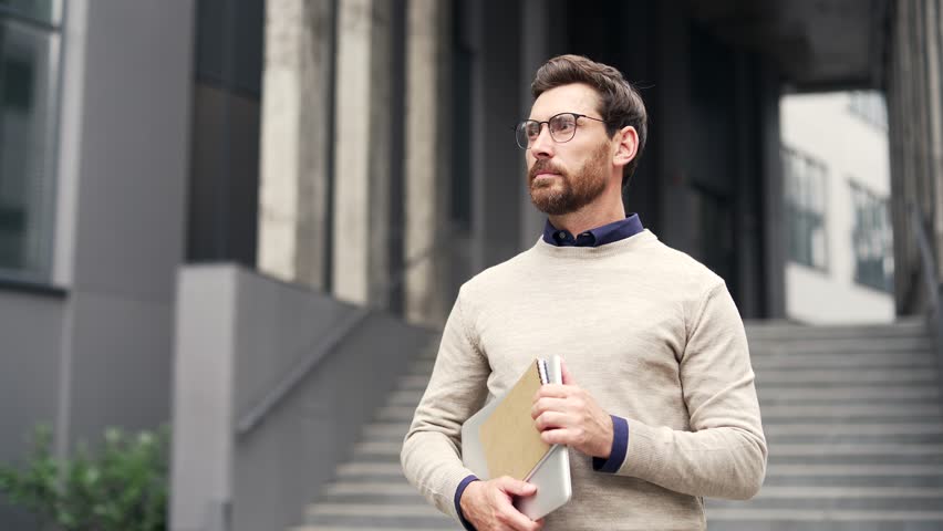 Smiling male businessman standing outside office building holding notebook and laptop. Head shot of happy mentor or teacher. Confident tutor posing and looking at camera. Friendly bearded man worker