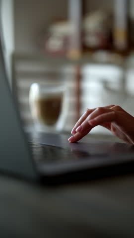 Vertical shot of unrecognizable businesswoman typing on laptop computer drinking coffee. Successful freelancer engaging in remote working from cozy living room at home, enjoying moment of relaxation.