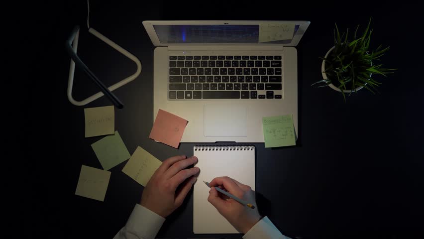 Overhead Shot of a Businessman in the Evening, Sitting at a Table and Sketching Time and Money Diagrams in a Notebook, A view from above showing a businessman working late into the evening
