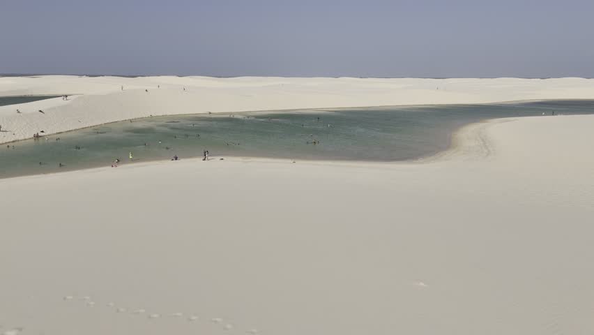 Drone flies low over sand dunes and over lake with people swimming in the Lençóis Maranhenses