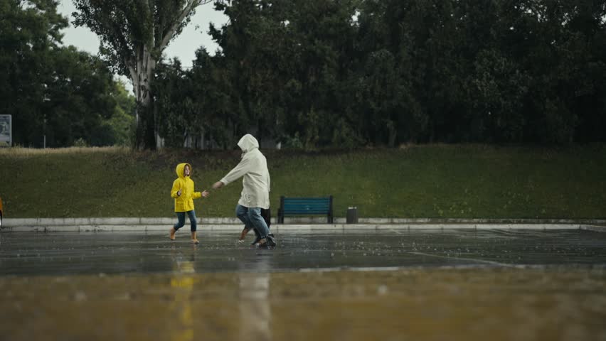 Side view of a happy teenage girl in a yellow jacket playing with her mom and a black dog during the rain in the park