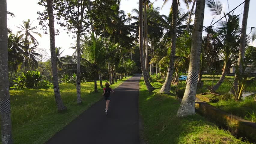 A young girl with a backpack, wearing denim shorts, runs along an atmospheric road illuminated by sunlight, along the edges of which palm trees grow.