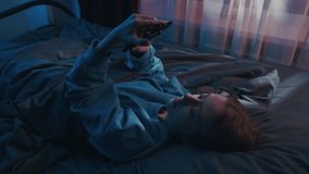A young woman lies on her back in bed, holding her phone and typing with a positive mood. It's early morning or night in her cozy bedroom, creating a warm, inviting atmosphere - Powered by Shutterstock - Get 15% off with code: PIKWIZARD15
