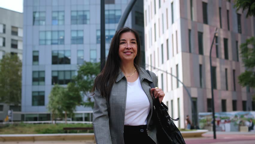Happy mature European business woman strolling confident to work with empowered expression outdoors. Professional adult urban female in formal attire with cheerful attitude walk to workplace in city
