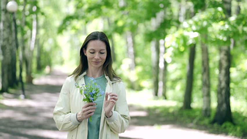 In the rays of daylight, among tall trees, a woman holds a bouquet of forget-me-nots, her face expresses calmness and joy from unity with nature.