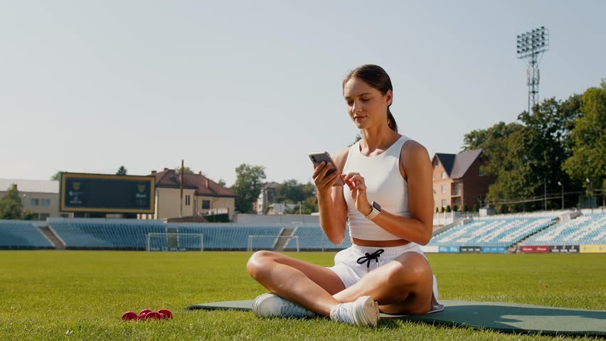 Smiling young woman using cell phone checking fitness applications sitting on stadium background outdoors. Online gym free exercises, sport and health care commercial apps concept.