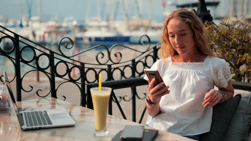 A woman with curly hair focuses on her smartphone while sipping a smoothie at a cafe by the water.