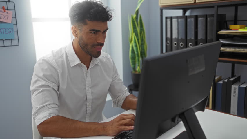 Young hispanic man working in an office setting, dressed in a white shirt, sitting at a desk with a computer, surrounded by shelves with binders, plants, and motivational notes on the board