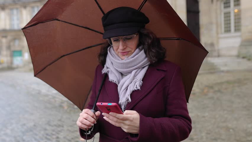 Young adult woman holding an umbrella during rainy day using an app on her mobile phone to find a location