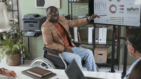 Full shot of young African American businessman in wheelchair presenting his new project to business team partners at conference, pointing at charts and diagrams on large digital monitor in office - Powered by Shutterstock - Get 15% off with code: PIKWIZARD15