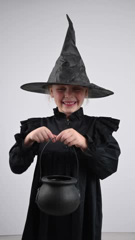 Portrait of a little Caucasian girl in a witch costume holding a cauldron on a white background. Trick o treat. Vertical video. 