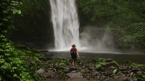 Solo traveler, a blonde woman with a backpack, approaching a majestic waterfall in a lush jungle. - Powered by Shutterstock - Get 15% off with code: PIKWIZARD15