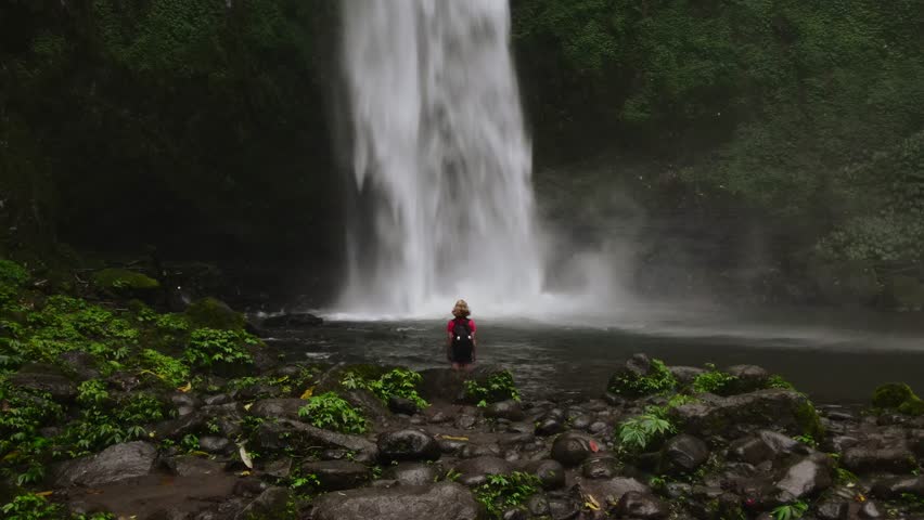 Solo traveler, a blonde woman with a backpack, approaching a majestic waterfall in a lush jungle.