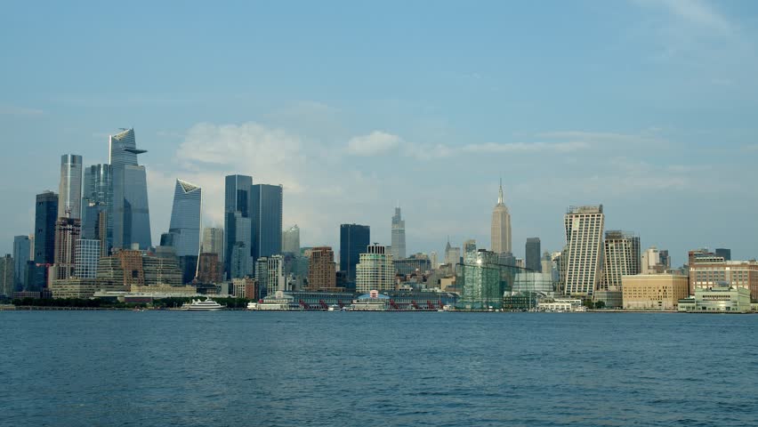 New York City skyline rises majestically across the river, towering skyscrapers standing tall against the sky. A few boats are scattered near the shore balanced by the peacefulness of the river
