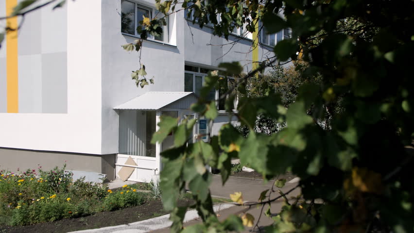 Entrance to multi-storey building with plastic doors. Courtyard with flowerbeds filled with different types of blooming flowers and lawns