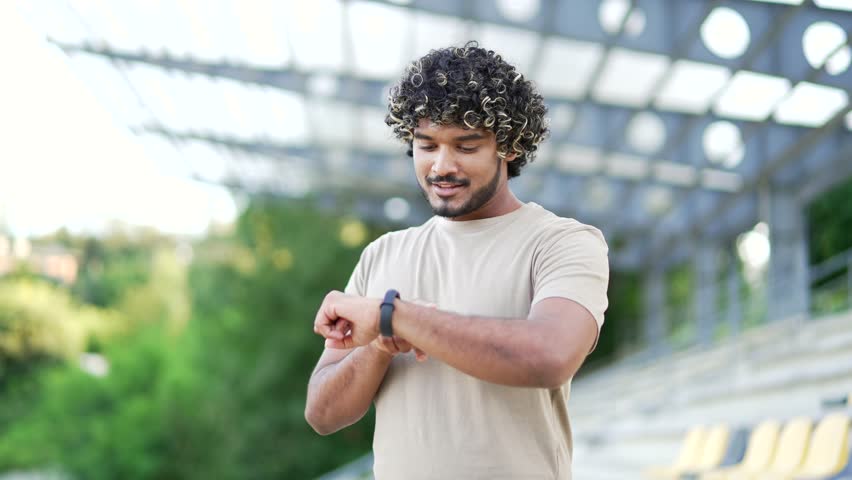 Smiling male checking smartwatch during outdoor workout. Celebrates success in fitness tracking. excitement and motivation while wearing casual t-shirt at modern sports venue after sports jogging
