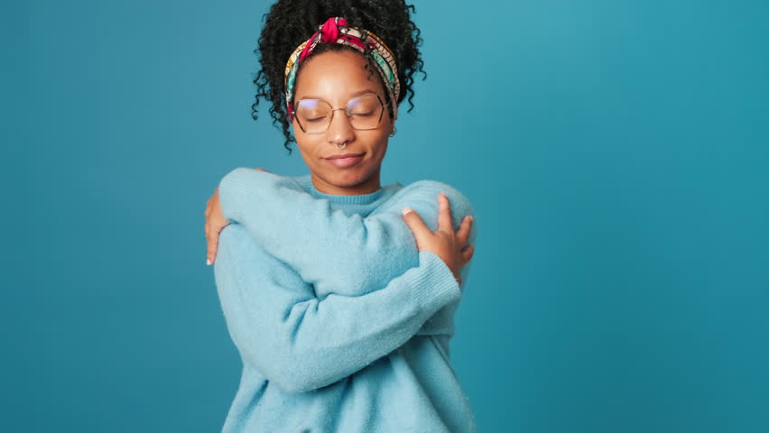 A smiling young woman with glasses, dressed in blue sweater, hugging herself with her arms, looks at camera, tenderness, on blue background in studio