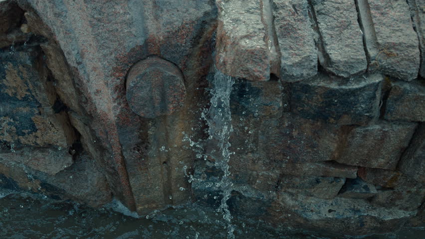 A stone wall with a small hole in it and water coming out of it. The water is falling in a steady stream