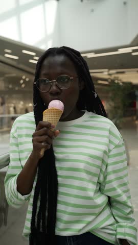 African american woman in striped shirt holding ice cream and experiencing tooth sensitivity, wearing striped shirt in indoor mall, discomfort while eating cold dessert