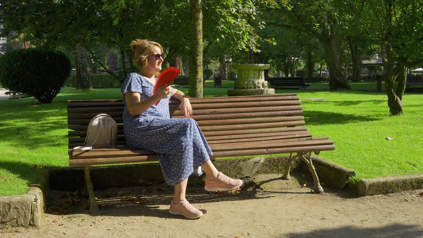 Woman sitting on a wooden bench in a public park and fanning herself on a hot day, Lugo, Galicia.