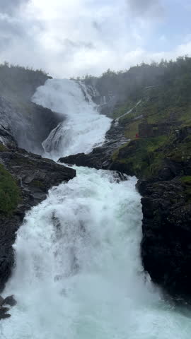 Vertical video, panning shot. Powerfull Kjosfossen waterfall in Western Norway, woman in red dress shows up from behind ruins.