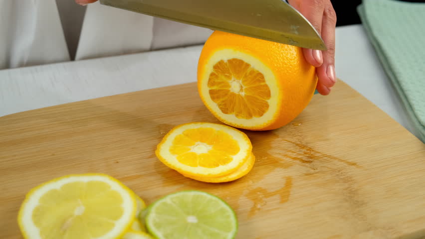 Woman cutting orange into slices using sharp knife on wooden kitchen board. Preparing lemonade from citrus fruit lemons, limes, oranges. Ingredients for homemade drink. Recipe cuisine culinary.
