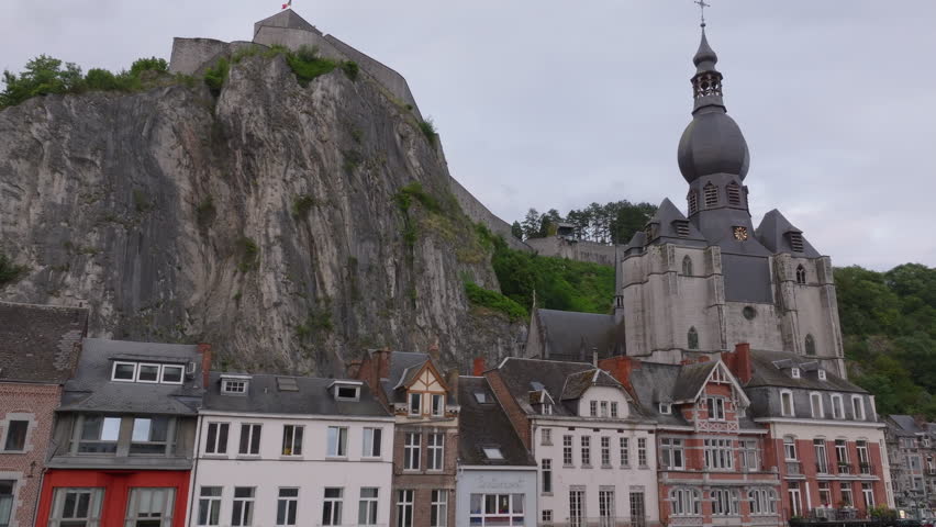 Houses line the banks of the Meuse river in Dinant, Belgium. A steep cliff towers over the homes with the Collegiate Church of Our Lady visible