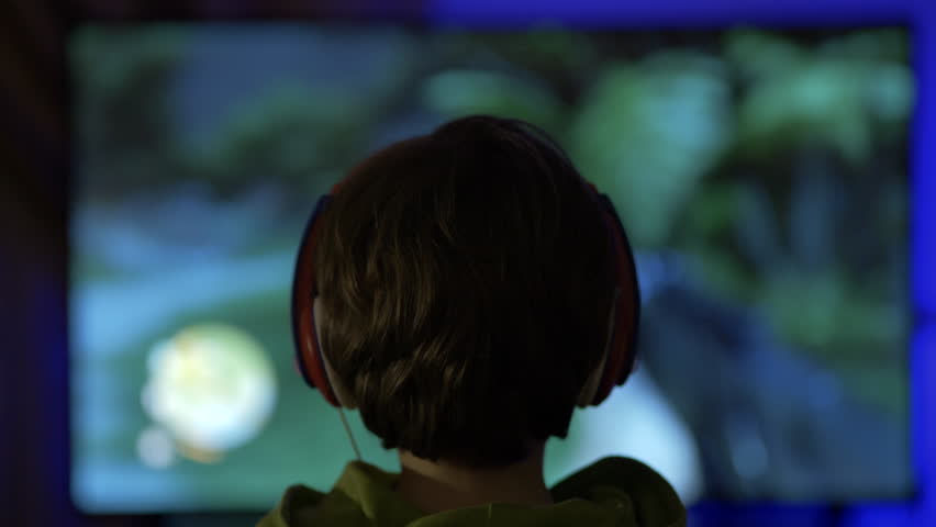 Child Gamer Playing Video Games on TV Screen. Boy in Headphones Playing Computer Video Game on Console at Night. Person in Headphones With Joystick Plays Video Game on Big TV screen. Online Games.
