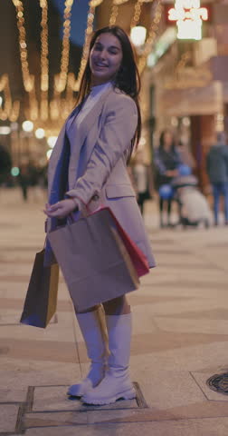 Pretty young brunette girl shopping around the city with gift bags and clothes at christmas, christmas lights, christmas decoration, vertical shot
