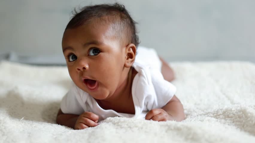 Happy laughing active healthy baby lying in bed trying to crawl waving hand at home. Portrait of adorable little kid having good mood. Rising son, first steps in crawling after hand plank, babycare.

