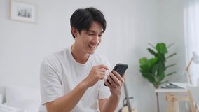 Asian excited man read online news on mobile phone in morning at home. Happy attractive male sitting on bed in bedroom enjoy holiday weekend, using smartphone communication with technology in house. - Powered by Shutterstock - Get 15% off with code: PIKWIZARD15